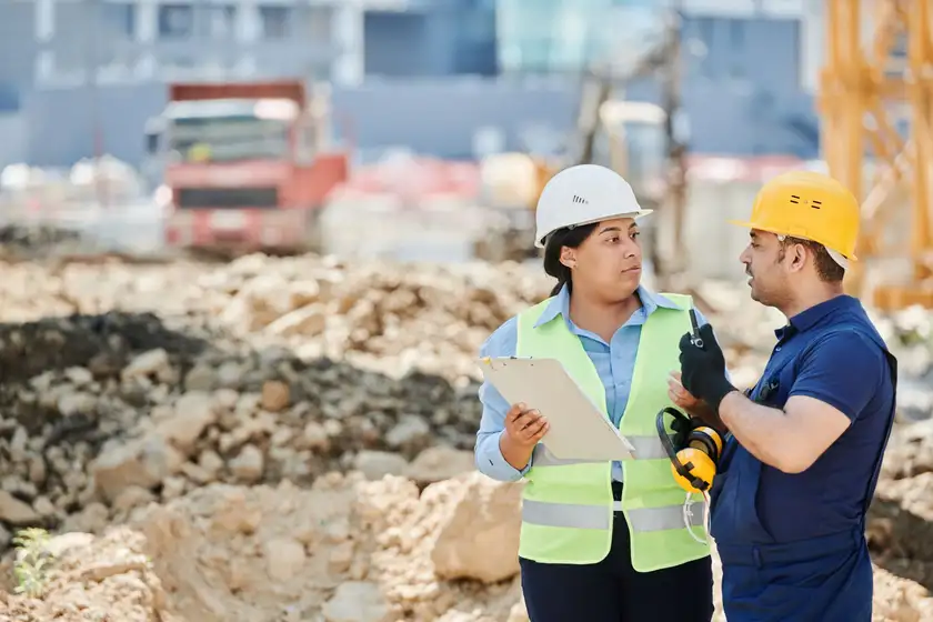 Two workers in hard hats and high-visibility vests reviewing plans at a construction site